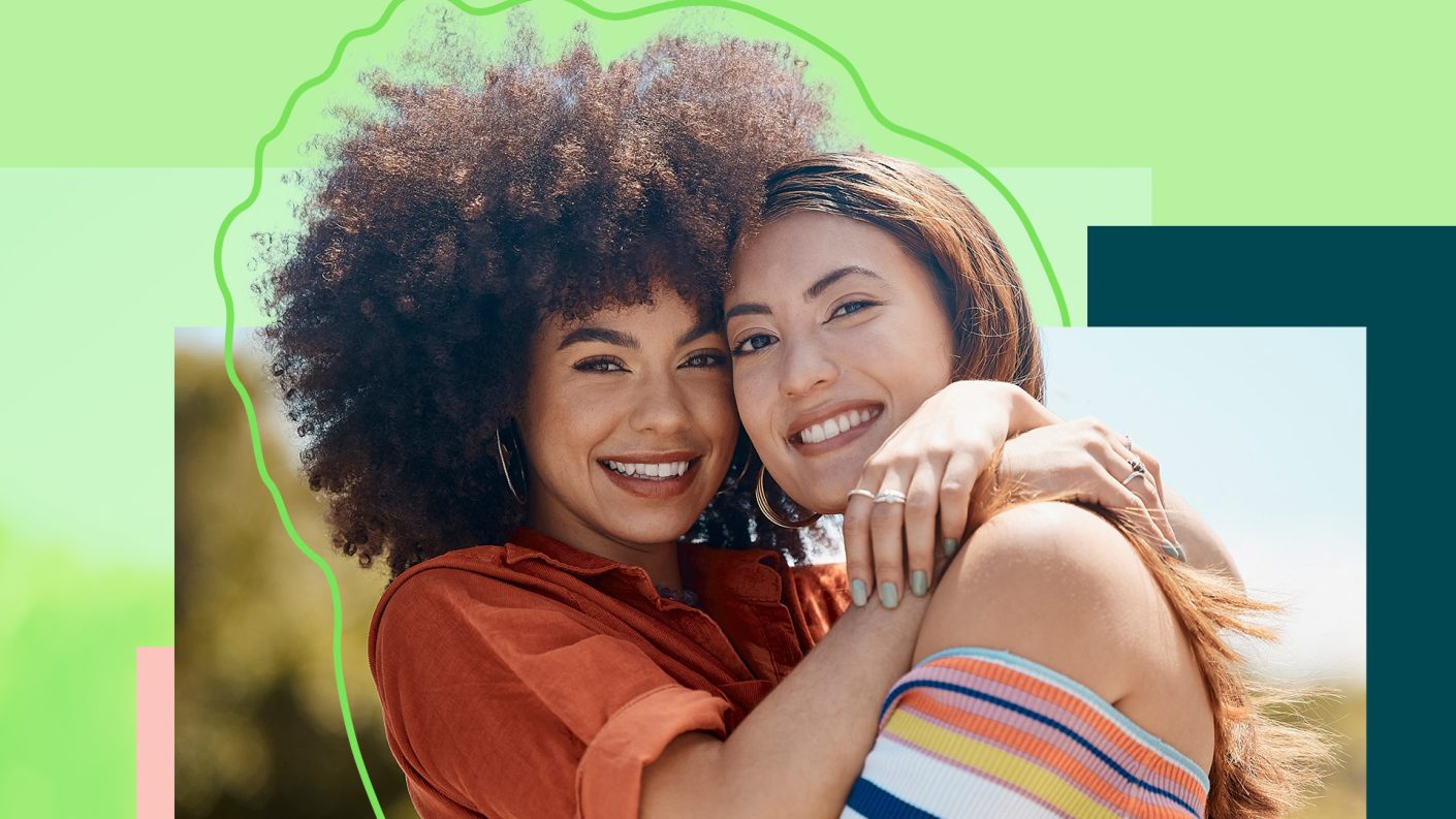 Two women smiling and hugging. The left woman has afro hair and wears an orange shirt. The right woman has Asian roots and wears a striped off-shoulder top.