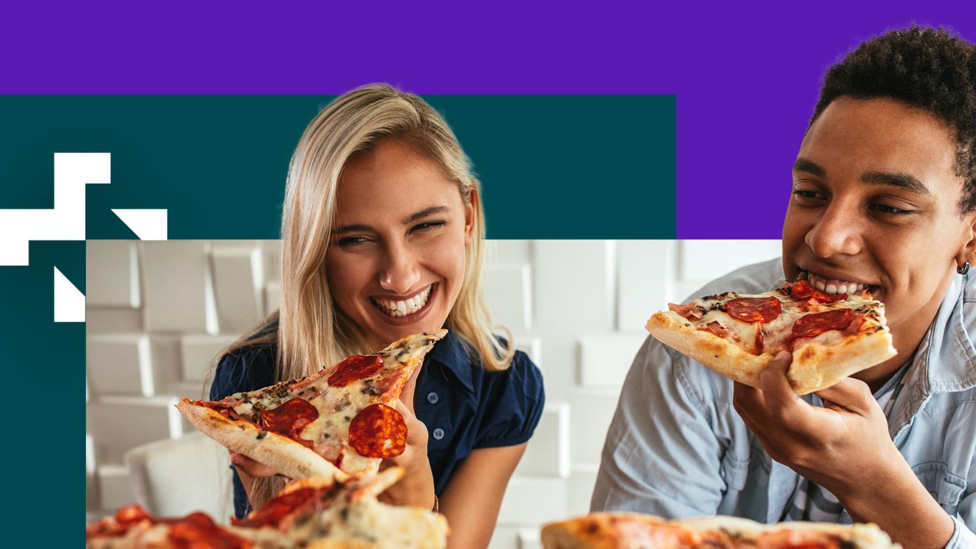 A white woman with blond hair and black man with black hair are smiling and eating pizza, sitting down at a table.
