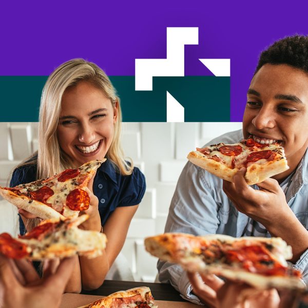 A white woman with blond hair and black man with black hair are smiling and eating pizza, sitting down at a table.